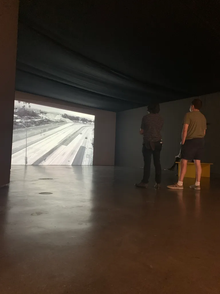 Photograph of two LA Art Show attendees watching Carmen Argote's film Last Light projected onto a gallery wall.