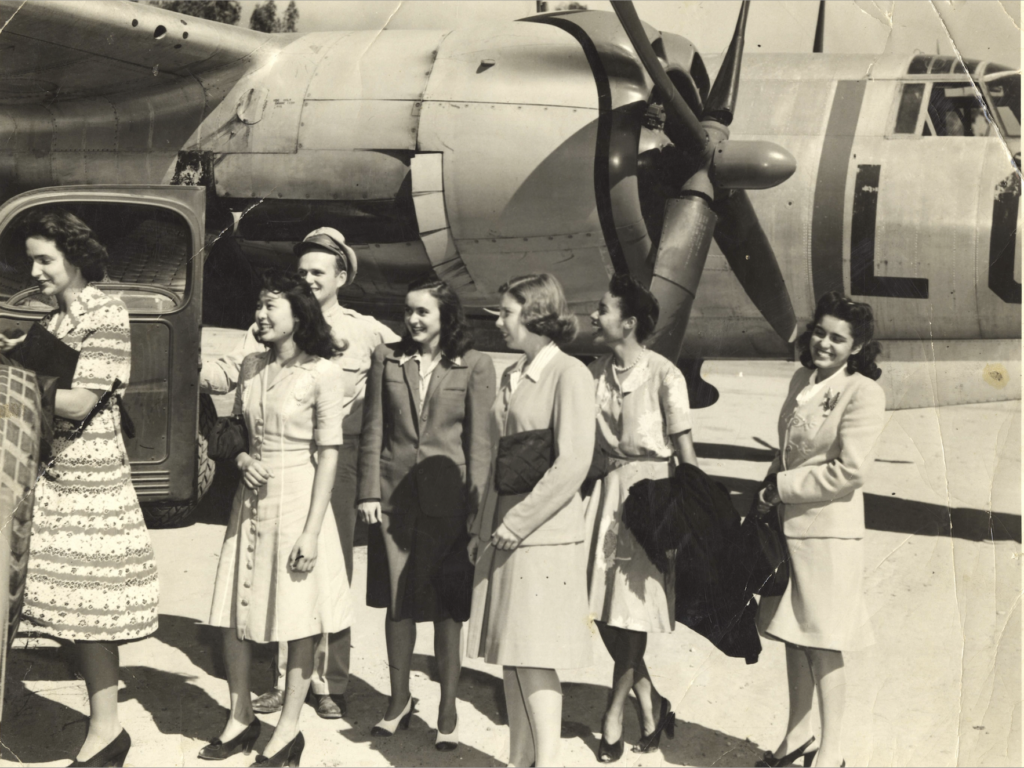 Black and white photo of seven members of the Panel of Americans standing in front of a military airplane in 1945.