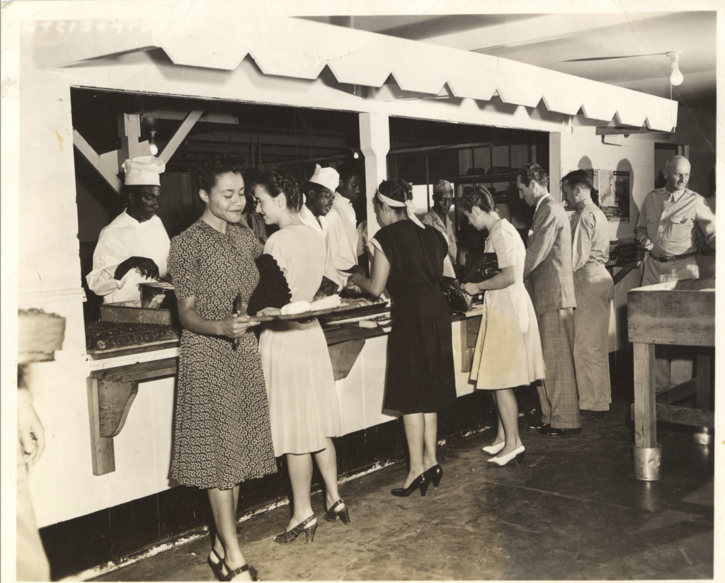 Black and white photo of seven Panel of Americans members visiting the mess hall at Fairfield-Suisun Army Air Base holding trays of food.