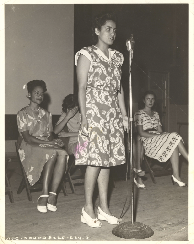 Black and white photo of a Panel of Americans member speaking at a microphone while three women sit behind her.