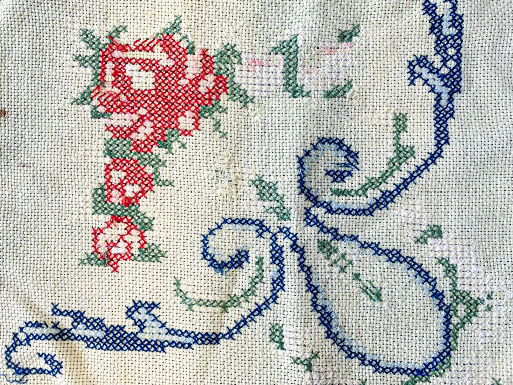 Close-up photograph of a cross-stitched white fabric with red flowers, green leaves, and blue decorative swirls.