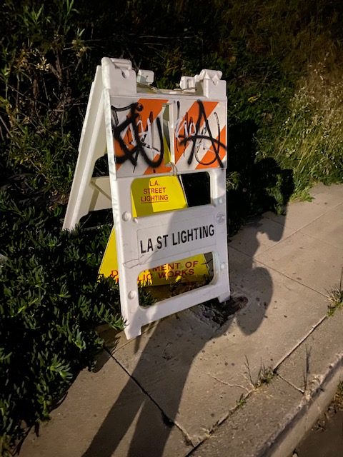 Photograph of a sidewalk sign along Carmen Argote's Last Light route, covered with graffiti with a traffic cone behind it.