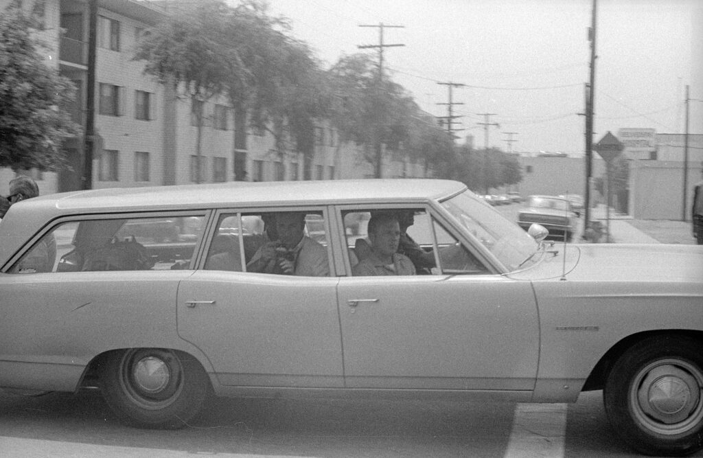 Black and white photo of four people sitting inside a car with tall buildings and trees behind the car.