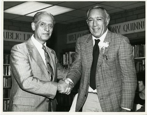 Black and white photo of Edward Roybal and Anthony Quinn, both wearing suits, smiling, and shaking hands inside a library.