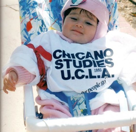 Photo of a baby wearing a white Chicano Studies UCLA shirt and a pink hat while sitting in a stroller.