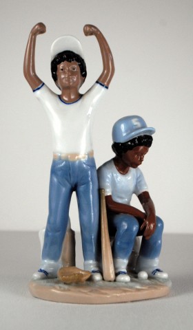 Porcelain sculpture of two young male baseball players in white t-shirts, blue pants and caps, one standing with both arms raised and one sitting.