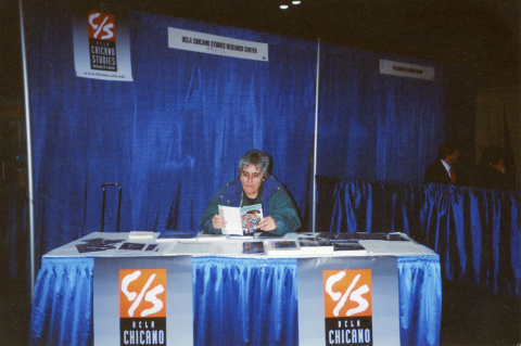 Yolanda Retter seated at a UCLA Chicano Studies Research Center information table at a conference, holding a booklet with blue curtains behind her.