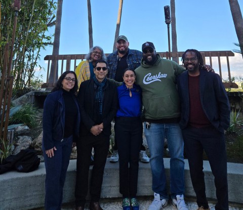 Seven people posing together outdoors in two rows, with trees and a wooden structure in the background.