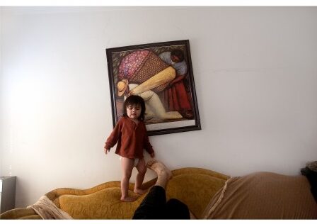 Photo of a young girl standing on a yellow couch in a living room with a framed painting hanging on the wall behind her.