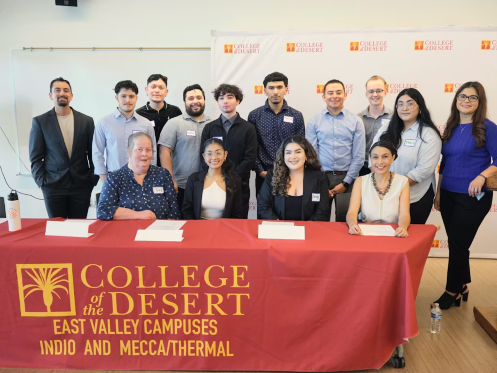 Photo of fourteen people posing together, some standing and others sitting at table with a red College of the Dessert tablecloth.