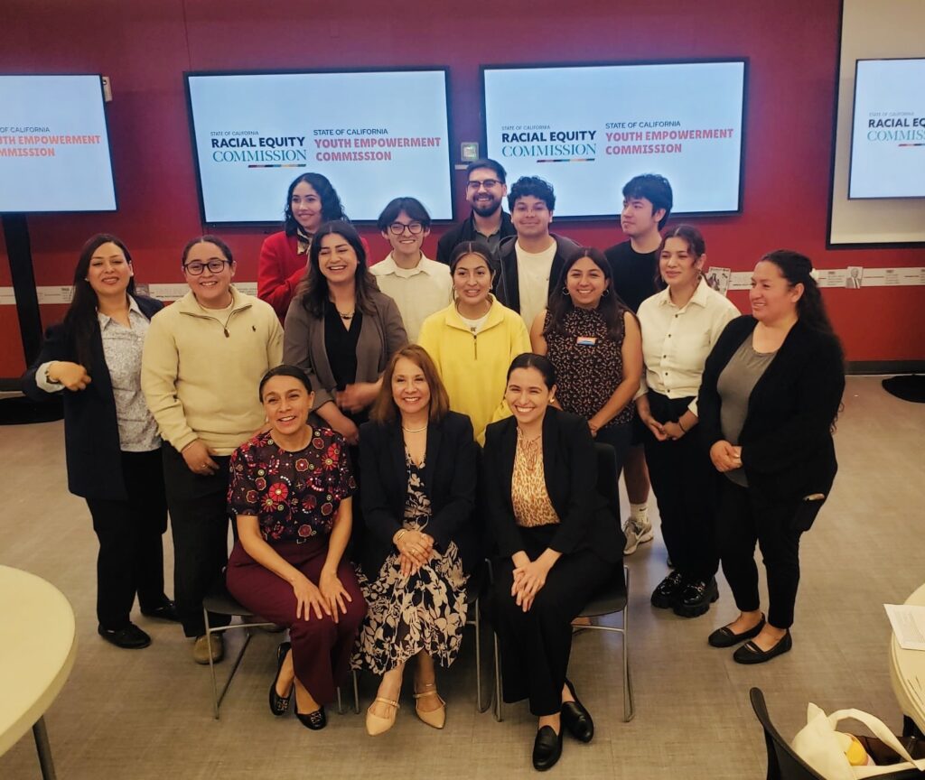 Photo of fifteen youth researchers, leaders, and representatives posing together in front of Racial Equity and Youth Empowerment Commission backdrops.