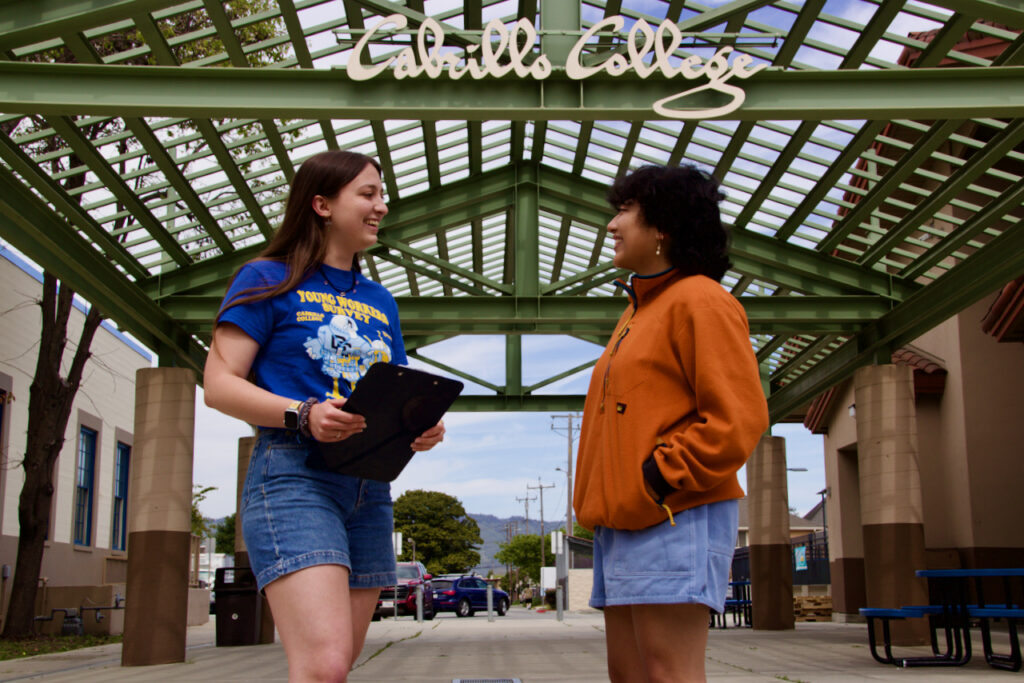 Photo of a youth researcher holding a clipboard while talking with a young person outside the Cabrillo College entrance.